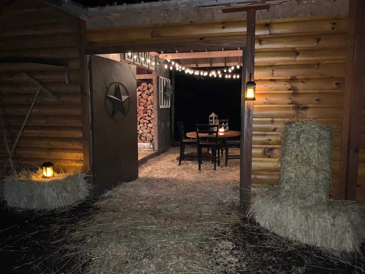 A rustic wooden barn setup with a star-accented door, dining table and chairs, string lights overhead, hay bales, and lanterns. The space feels warm and is styled for memorable group gatherings and celebrations under the stars.