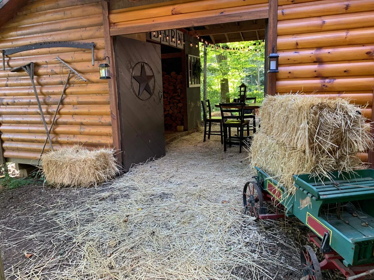 An inviting exterior scene showing a wooden cabin flanked by straw bales, rustic farm equipment, and a dining table set up for outdoor meals. The open-air setting suggests communal gatherings and evenings beneath the stars.