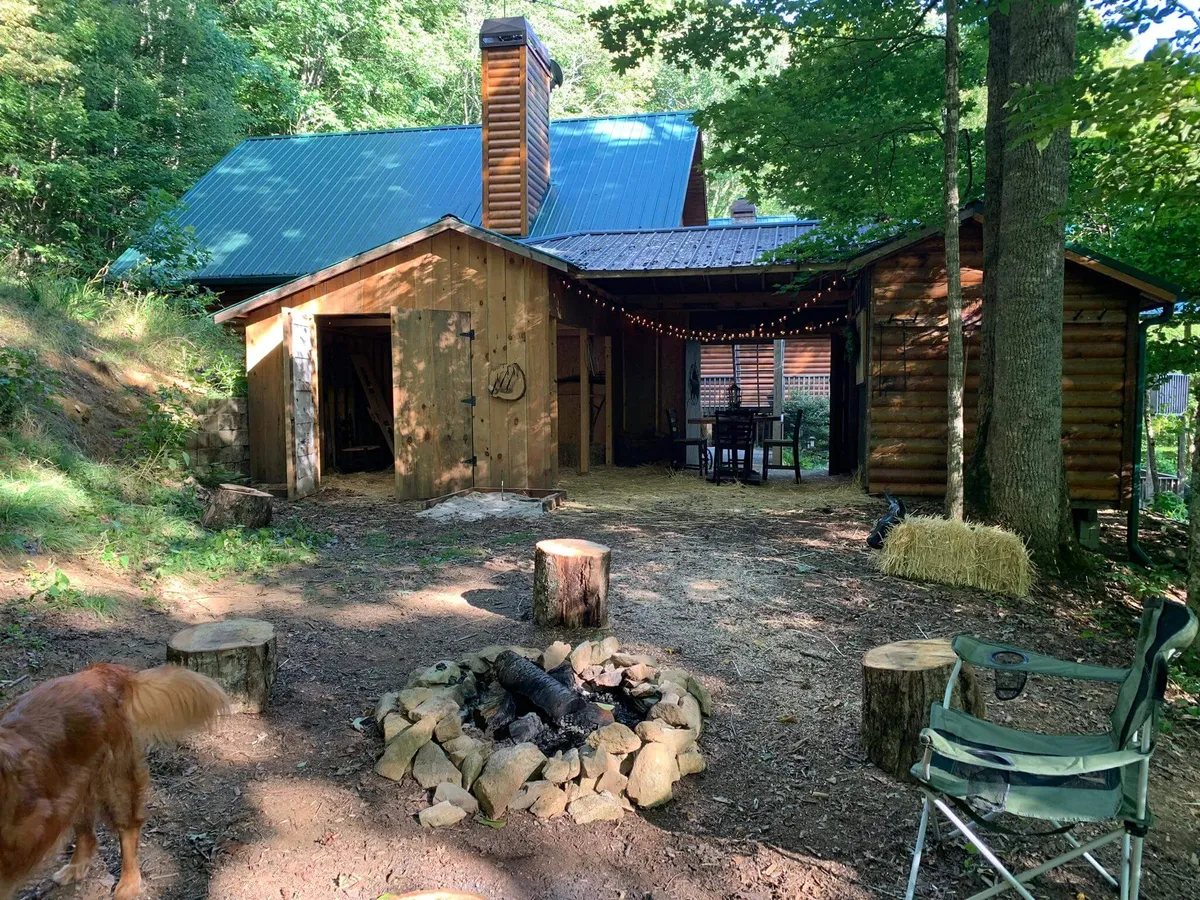 An inviting fire pit area with a wooden cabin backdrop, outdoor dining table, and surrounding trees. The setup is ideal for campfire gatherings, outdoor dining, sharing stories, and enjoying s’mores beneath the canopy of nature.