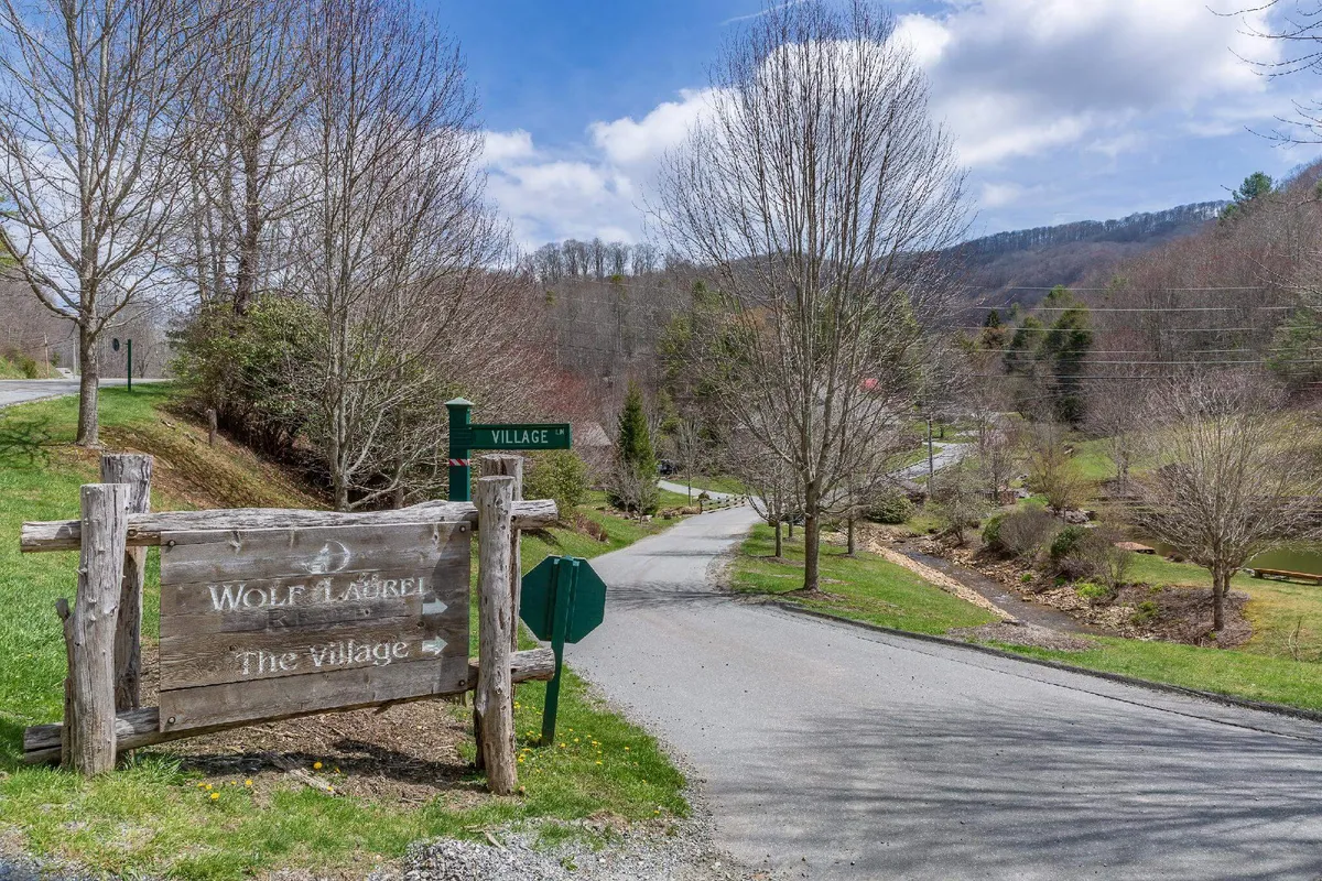 A charming wooden sign marks the entrance to a village, with a road leading into a wooded area. The surrounding trees and community setting evoke a sense of exploration, discovery, and access to local amenities or hidden trails.