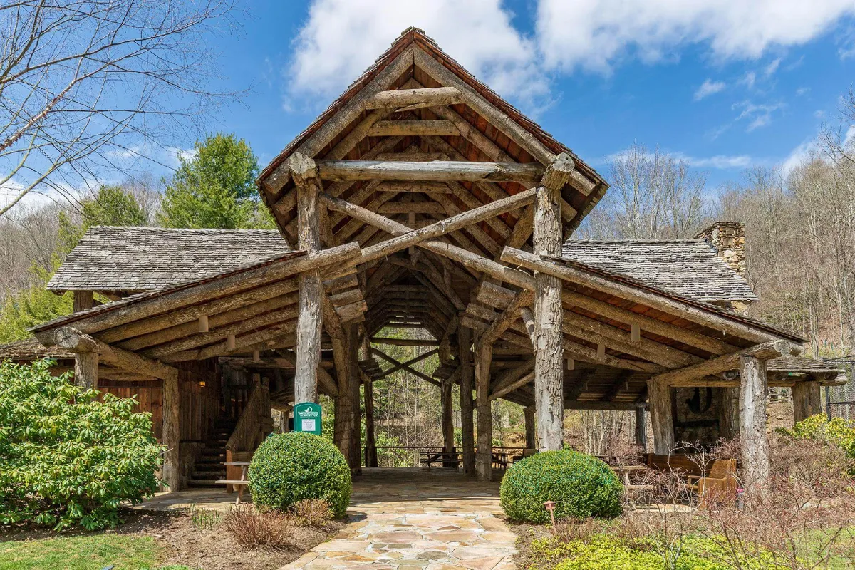 A rustic wooden structure surrounded by lush greenery, with a stone pathway leading up to the porch. The scene is surrounded by trees and natural landscaping, evoking a sense of adventure and connection to the outdoors. Architectural details of the structure are visible, and the inviting porch could be a spot to enjoy nature or set out for a hike.