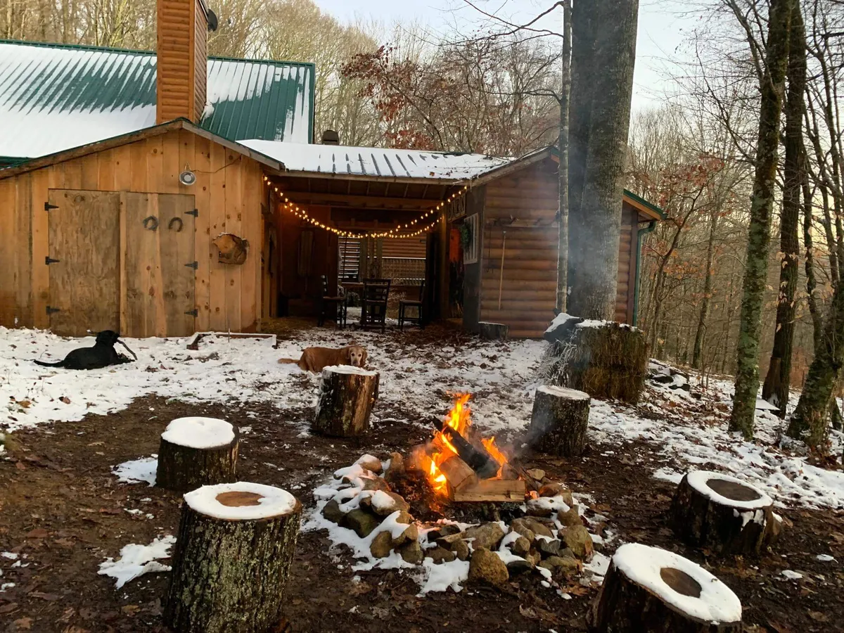 A cozy campfire scene with wooden seating logs encircling the fire, string lights overhead, and a log cabin in the background. The image is set outdoors and suggests activities like gathering around the fire, storytelling, stargazing, and celebrating holidays with loved ones.