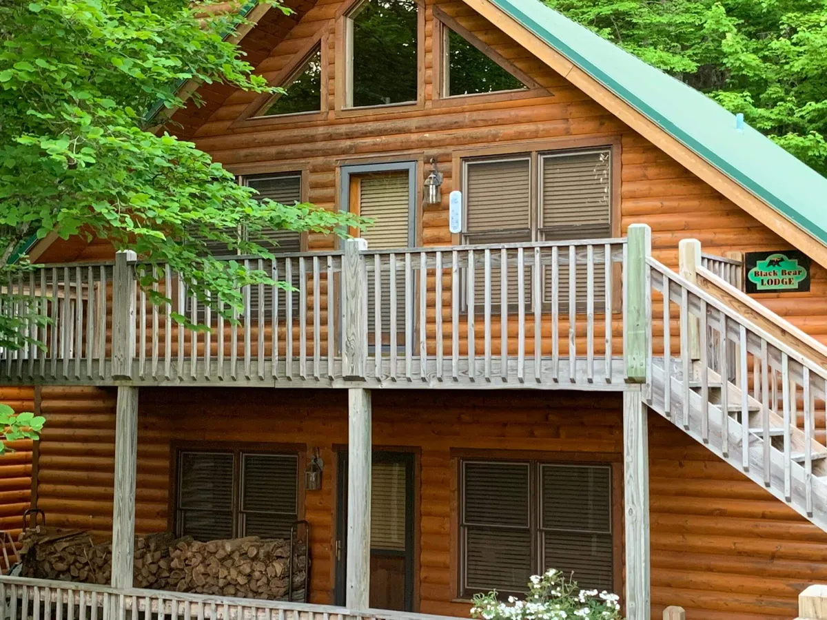 The image showcases a log cabin structure with a green roof and wooden balcony railings. A sign reading 'Black Bear Lodge' is visible, and stacked logs sit below the balcony. The setting is surrounded by natural scenery, giving a sense of privacy and serenity. The image hints at activities such as relaxing on the balcony, barbecuing outdoors, and stargazing at night.