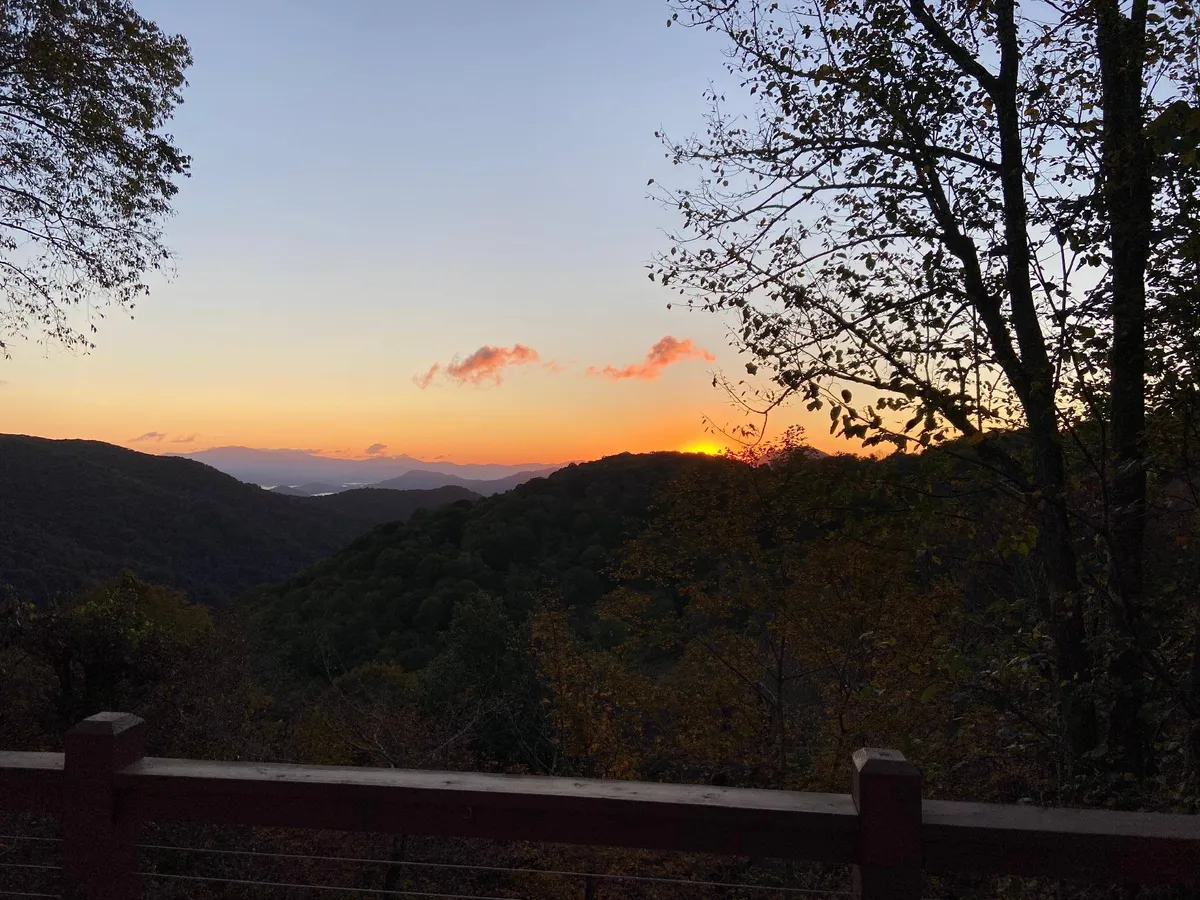 A sweeping mountain view at sunset, with a vibrant sky framing the silhouette of the mountain range and trees in the foreground. The atmosphere is tranquil, inviting, and awe-inspiring—ideal for hiking, nature walks, or simply soaking in the scenery.