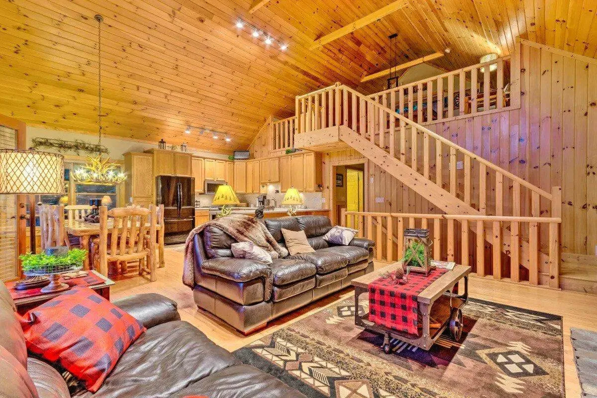 A wide, inviting view of the main living area featuring a cozy leather sofa, a warm wooden staircase, a large dining table, and a kitchen area in the background. The space is filled with natural light and has a rug that adds to the warmth and comfort of the room. This image captures the open-plan nature of the main cabin, perfect for gatherings and shared meals.