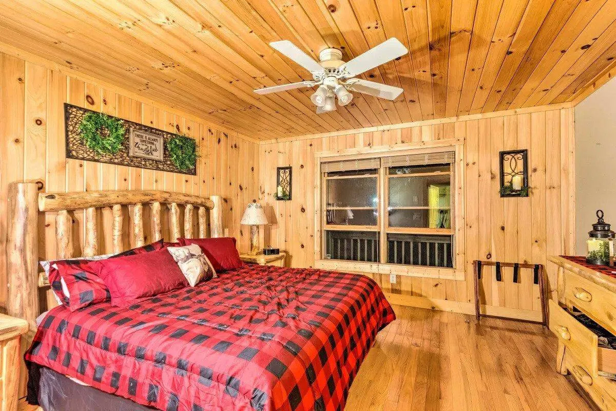 A warmly lit guest bedroom featuring a rustic wooden log bed adorned with plaid bedding, wood-paneled walls, and a window letting in soft daylight. The setting is quiet and inviting, designed for relaxation and privacy.
