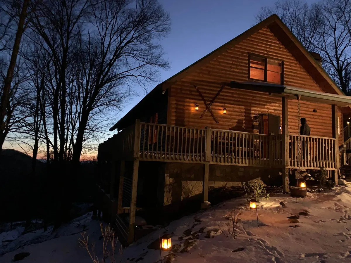 A charming log cabin stands among snowy trees, its spacious deck adorned with glowing lanterns inviting guests to enjoy the mountain air and tranquil winter views. The scene exudes warmth and serenity, highlighting the perfect setting for evening gatherings and peaceful relaxation in a cozy mountain retreat.