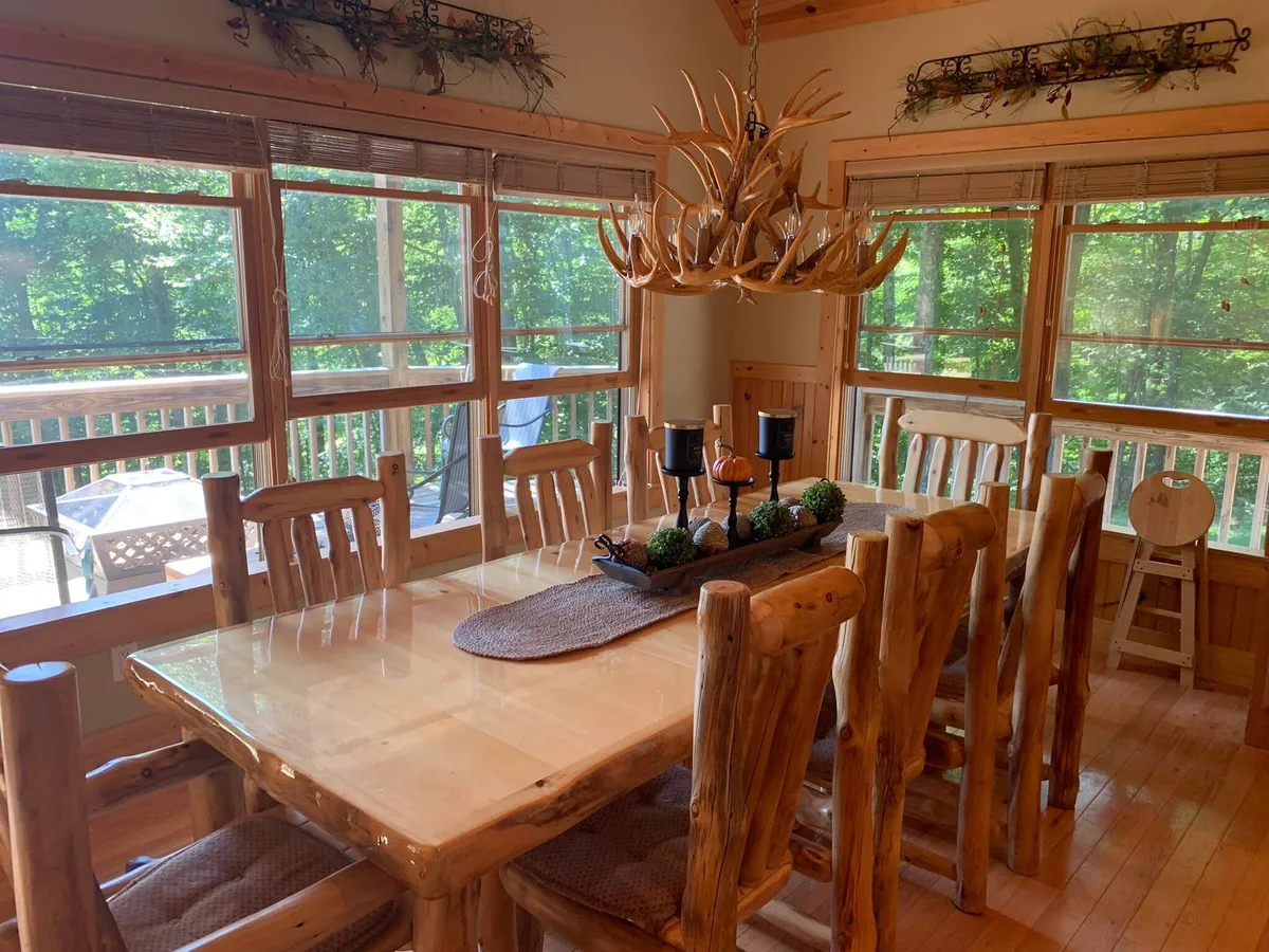 A large wooden dining table sits under an antler chandelier, flanked by large windows offering a forest view. The table is set with decorative centerpieces, inviting guests for memorable meals. The soft lighting and natural surroundings outside the windows create a cozy and welcoming atmosphere.