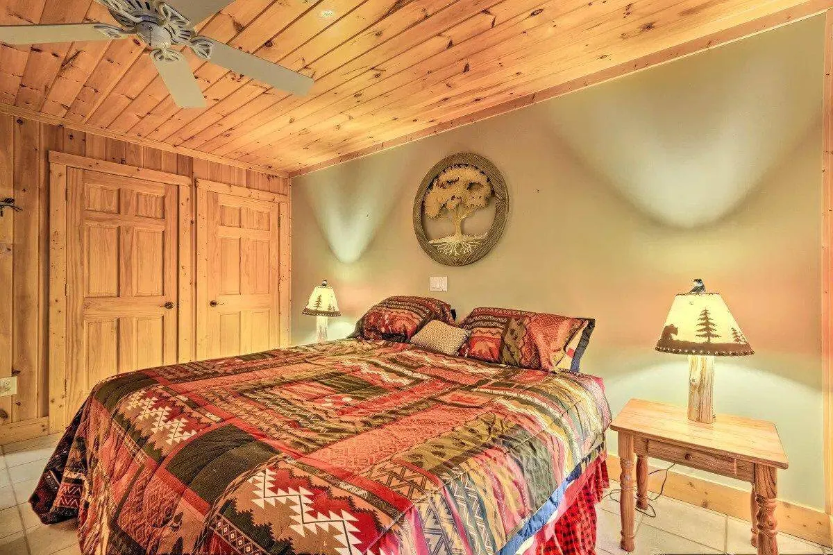 A spacious bedroom with a bed dressed in a patterned quilt, flanked by nightstands with warm-glowing lamps. Wooden closet doors and decorative wall art complete the comfortable scene, with sunlight gently filtering in.